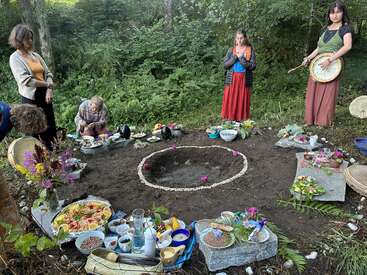 Cinco personas se reúnen al aire libre alrededor de un altar circular de tierra decorado con flores, piedras, comida y ofrendas, y parecen estar participando en un ritual espiritual o ceremonial.