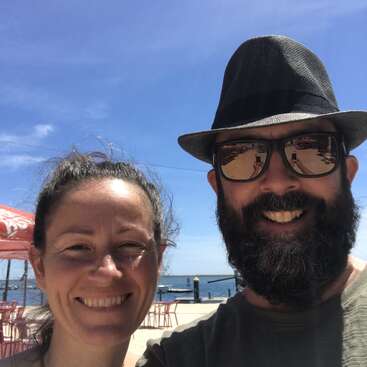 Eine lächelnde Frau und ein bärtiger Mann mit Sonnenbrille und Hut machen ein Selfie am Wasser unter einem strahlend blauen Himmel.