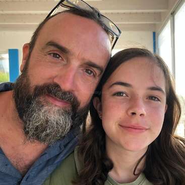 A bearded man and a young girl smile closely together in a sunlit indoor setting, with large windows behind them, creating a warm, happy atmosphere.