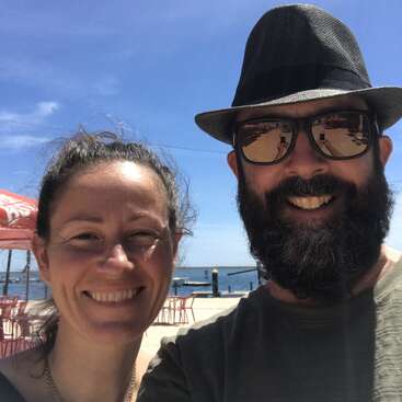 Uma mulher sorridente e um homem barbudo usando óculos escuros e chapéu tiram uma selfie ao ar livre perto da água sob um céu azul brilhante e ensolarado.