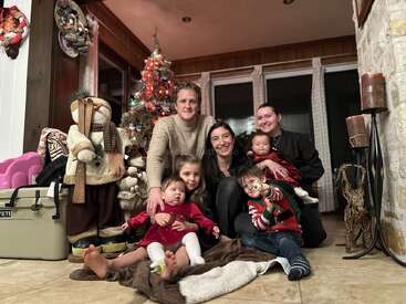 Une famille pose joyeusement dans un salon confortable décoré pour Noël, avec un sapin illuminé, des figurines festives et des enfants vêtus de tenues de fête, souriant chaleureusement.