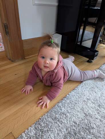 Un bébé avec une queue de cheval, vêtu d'une tenue mauve, rampe sur un sol en bois à côté d'un tapis gris, levant les yeux avec un regard vif et curieux.