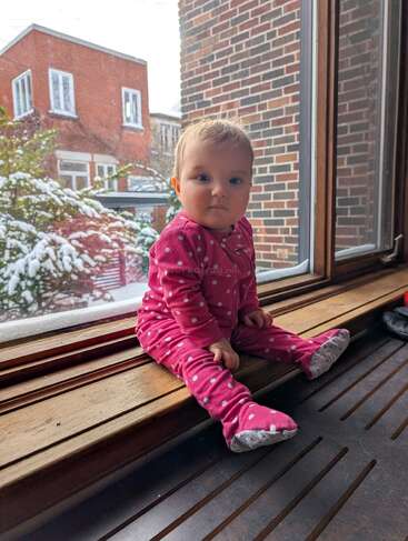 Un bébé vêtu d'une grenouillère rose à pois est assis sur un rebord de fenêtre en bois, contemplant les arbres enneigés et les bâtiments en briques rouges par une journée d'hiver.