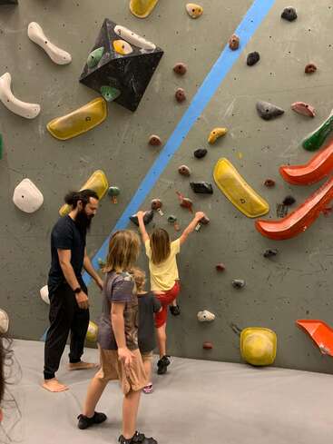Trois enfants et un adulte se trouvent dans une salle d'escalade. Un enfant escalade le mur coloré pendant que les autres le regardent et que l'adulte le surveille de près.
