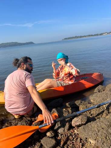 Un homme et une femme se trouvent au bord d'un lac calme et se préparent à faire du kayak. La femme est assise dans un kayak orange et porte un foulard bleu, tandis que l'homme l'aide.