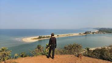 Une personne vêtue de noir se tient sur une falaise, surplombant une plage sereine, un océan bleu, un banc de sable et une végétation luxuriante sous un ciel bleu clair. Scène paisible.