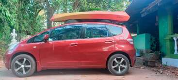 Une voiture rouge est garée à l'extérieur, à côté d'une maison verte. Un kayak orange vif est fixé sur le toit de la voiture. Des arbres et des plantes entourent la scène.