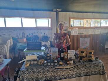 Uma mulher está atrás de uma mesa decorada com produtos caseiros, como potes, garrafas e frutas e legumes, em uma banca de mercado dentro de uma sala bem iluminada.