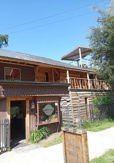 Sous un ciel bleu limpide, un bâtiment rustique en bois portant l'enseigne « Willhouse Hostel » se dresse au milieu de la verdure et des arbres, doté d'un porche accueillant et d'une terrasse surélevée.
