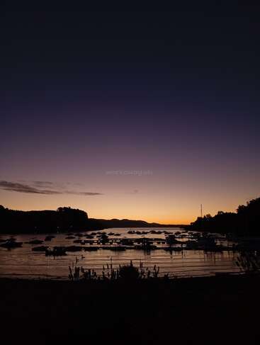 Un coucher de soleil paisible sur un lac tranquille, des bateaux flottant doucement sur l'eau, des arbres et des montagnes se découpant en silhouette à l'arrière-plan, le tout sous un magnifique ciel aux teintes pourpres et orangées.