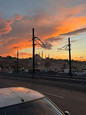 La imagen representa un paisaje urbano al atardecer, con un puente con coches, líneas eléctricas y edificios, sobre un cielo naranja y amarillo con nubes.