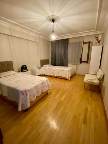 Esta imagen muestra un dormitorio ordenado con dos camas individuales, colchas de flores, suelos de madera, una mesilla de noche, iluminación moderna, una silla y un armario.