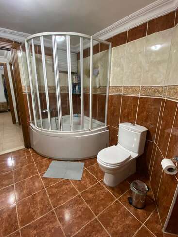 Este cuarto de baño presenta paredes y suelo alicatados en marrón y crema, una cabina de ducha curva de cristal, un inodoro moderno, una papelera plateada, un portarrollos de papel higiénico y una alfombrilla.