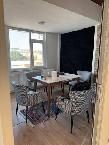 Una habitación luminosa y acogedora con cuatro sillas a cuadros, una mesa de madera con un portátil, una alfombra de colores, grandes ventanales y un panel negro insonorizado en la pared.
