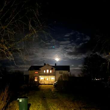 A house stands illuminated at night, surrounded by trees and darkness. The moon glows through dramatic clouds, casting a mysterious, serene atmosphere over the quiet scene.