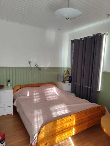 This is a cozy bedroom with a wooden bed, light pink blanket, white walls, green wainscoting, grey curtains, bedside tables, and a ceiling lamp. Sunlight streams in.