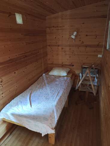 This small, cozy wooden room features a single bed with a light blanket and pillow, a wall lamp, and a compact side table or wooden stand.