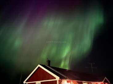 A mesmerizing green aurora lights up the night sky above a red-roofed house, creating a magical, serene, and breathtaking scene in the wilderness.