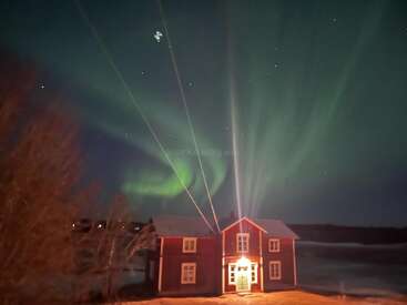 A cozy red house glows warmly under a spectacular night sky, illuminated by vibrant green Northern Lights and stars, creating a magical, serene, and enchanting winter scene.