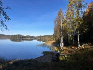A peaceful lakeside scene with calm water reflects a clear blue sky. Trees with autumn foliage edge the shore. Wooden benches and a dock invite relaxation.