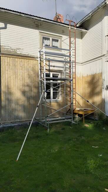 A wooden house exterior is being renovated. Scaffolding is set up by a window, with a ladder nearby. Patches reveal scraped paint. Grass covers the ground.