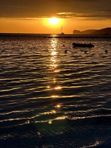Um pôr do sol dourado lança uma luz quente sobre a água suavemente ondulada. Barcos em silhueta descansam pacificamente, enquanto penhascos distantes completam essa cena tranquila e bela de paisagem marinha.