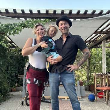 Eine lächelnde Familie steht im Freien unter einer Pergola. Die Frau und der Mann halten freudig ein kleines Kind. Grünpflanzen und Gartenmöbel schmücken die sonnige, entspannte Umgebung.