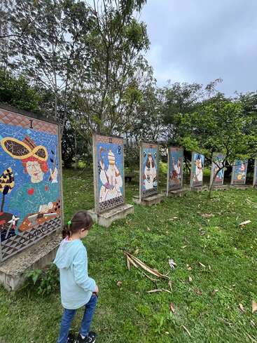 A young girl walks on grass, observing colorful mosaic panels depicting artistic figures outdoors. Trees surround the area under a cloudy sky, creating a peaceful, contemplative atmosphere.