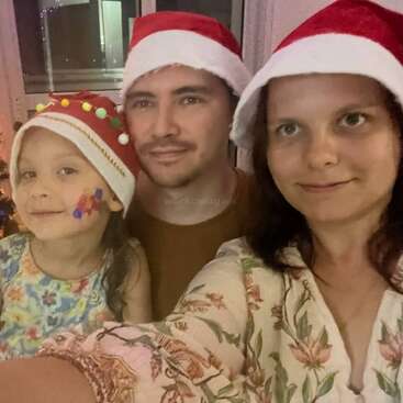 A family of three, wearing red Santa hats, poses together for a festive holiday photo. The child has face paint and all are smiling warmly.