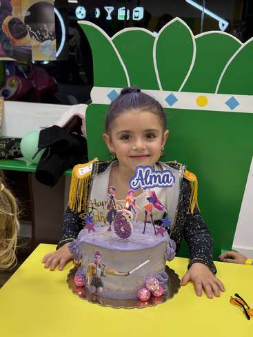 Una joven con el pelo morado está sentada sonriendo detrás de una tarta de cumpleaños decorada con personajes animados y su nombre, «Alma», lo que indica que se trata de la celebración de su cumpleaños.