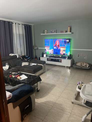 La foto muestra una acogedora sala de estar con un sofá modular, un televisor en el que se ve un programa, una mesa de centro con aperitivos, un perro en una cama y un andador para bebés.