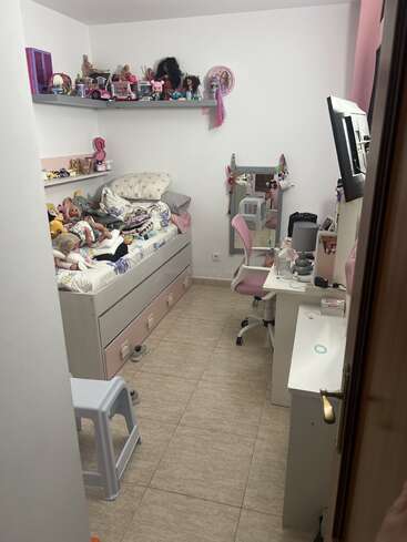 Una habitación de niña acogedora y organizada con una cama, un escritorio y una silla rosa. Las muñecas y los juguetes están expuestos en estanterías. Hay un espejo y un pequeño taburete.