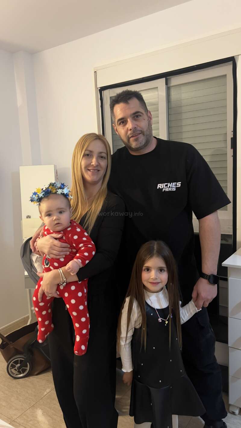 Una familia de cuatro miembros posa en el interior. La madre sostiene a un bebé vestido con un traje rojo de lunares, el padre está de pie a su lado y una niña sonriente está delante.