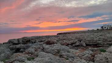 Una espectacular puesta de sol llena el cielo de tonos rosas y naranjas sobre una costa rocosa. El tranquilo mar y una casa lejana completan la serena escena.