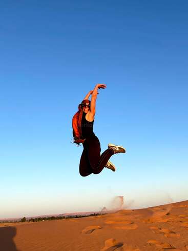Una persona salta alegremente en el aire sobre dunas de arena, con gafas de sol y un pañuelo rojo, bajo un inmenso cielo azul despejado durante la puesta o la salida del sol.