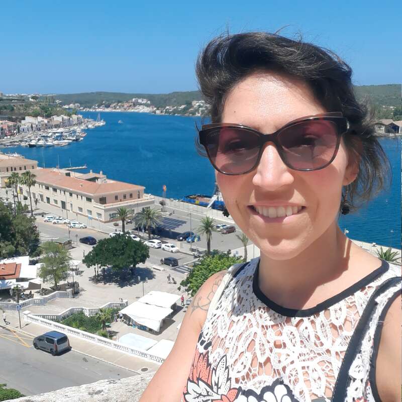 Una mujer con gafas de sol sonríe para hacerse un selfie con una preciosa ciudad costera, agua azul, barcos y edificios blancos bajo un cielo despejado y soleado de fondo.