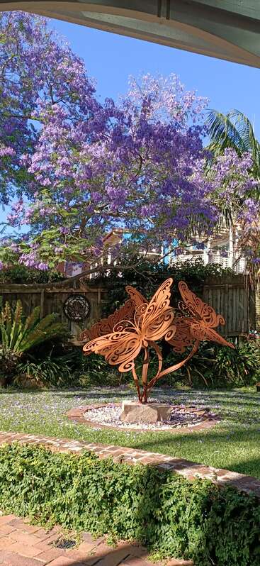 A vibrant garden features a large jacaranda tree with purple blooms, lush greenery, and a striking metal butterfly sculpture shining in the sunny, serene yard.