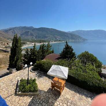 La imagen representa un sereno patio con una sombrilla blanca, rodeado de exuberante vegetación y con vistas a una tranquila masa de agua y a majestuosas montañas bajo un cielo azul despejado.