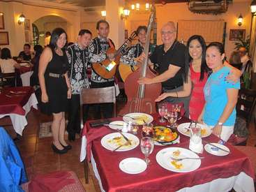Das Bild zeigt eine Gruppe von Menschen, die für ein Foto in einem Restaurant posieren, mit einem für das Abendessen gedeckten Tisch und einer Mariachi-Band im Hintergrund.