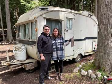 Ein lächelndes Paar steht gemeinsam vor einem Oldtimer-Wohnwagen auf einem bewaldeten Campingplatz, umgeben von hohen Bäumen und der Natur, und genießt die Natur.