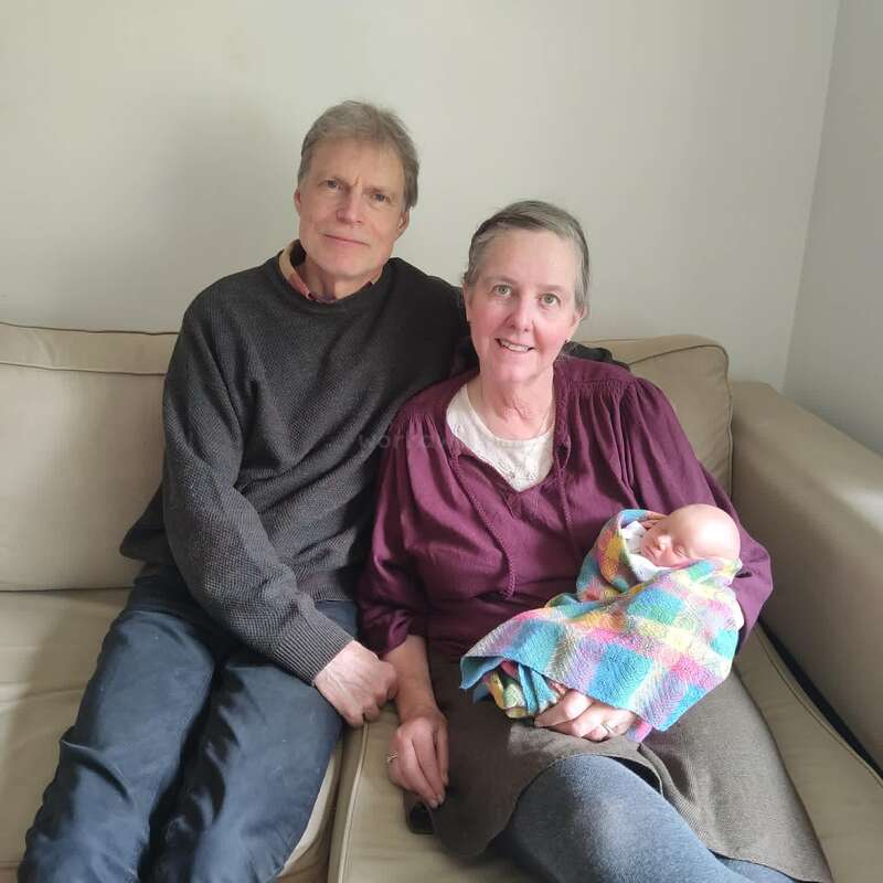 Ein älterer Mann und eine ältere Frau sitzen eng beieinander auf einem beigen Sofa. Die Frau hält liebevoll ein schlafendes Neugeborenes im Arm, das in eine bunte Decke gewickelt ist. Alles wirkt friedlich.