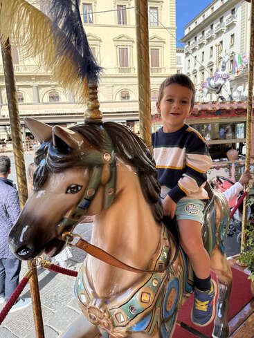 Un niño con camisa de rayas y pantalones cortos monta alegremente un caballo de carrusel decorado en una animada plaza europea iluminada por el sol y rodeada de edificios históricos.