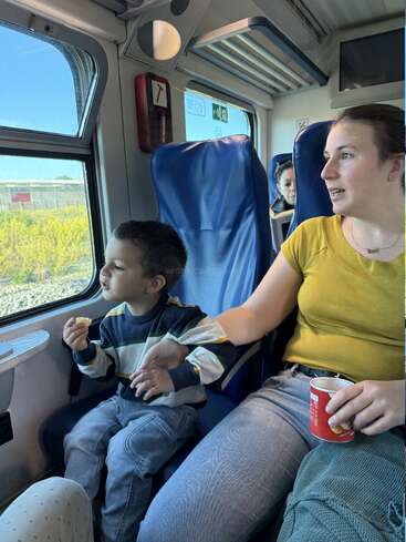 Una mujer y un niño se sientan juntos en un tren. El niño mira por la ventanilla, con un bocadillo en la mano, mientras la mujer sostiene una taza roja.