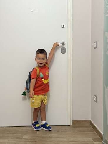 Un chico joven con una camiseta roja y pantalones cortos amarillos está de pie junto a una puerta blanca, sonriendo, con una mochila puesta y tendiendo la mano hacia el pomo de la puerta.