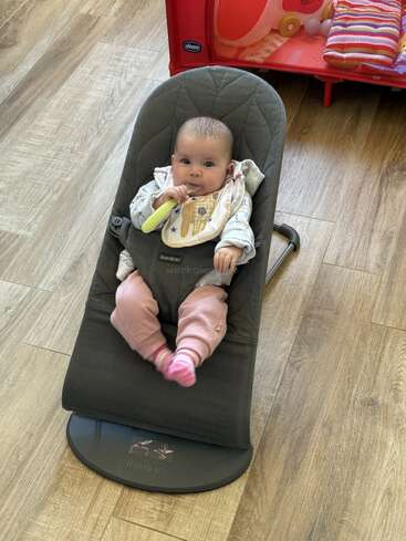 Un bebé está sentado en una hamaca BabyBjorn gris, sosteniendo una cuchara verde. El bebé lleva pantalones rosas, babero y calcetines a rayas, sobre un suelo de madera.