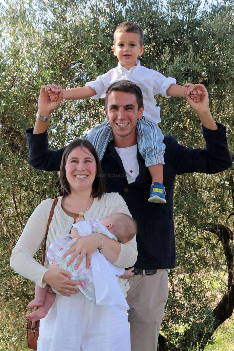 Una familia feliz posa al aire libre. El padre lleva a hombros a un niño pequeño, mientras que la madre, sonriente, sostiene a un bebé en brazos. Fondo de árboles.