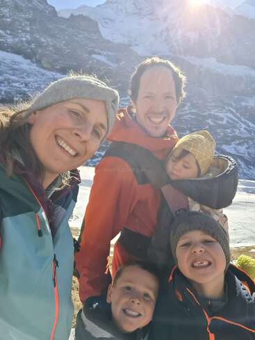 L'image représente une famille de cinq personnes posant pour une photo devant une chaîne de montagnes enneigées, le soleil brillant derrière eux.