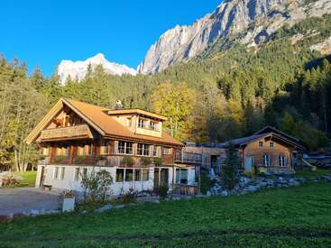 A charming wooden house sits surrounded by lush forest and bright greenery, with majestic mountains and clear blue skies in the background, evoking tranquility and beauty.