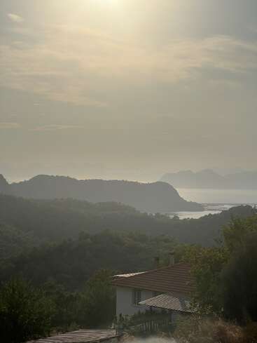 A serene landscape at sunrise, featuring a house in the foreground, lush green hills, distant mountains, and a misty lake under soft, golden light.