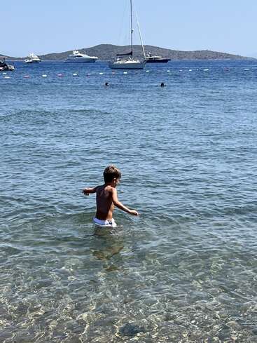 A young boy wades in clear blue sea water, near the shore. In the distance, people swim and several yachts and a sailboat are anchored.